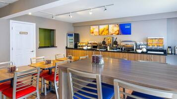 A modern hotel room with a dining area featuring a long wooden table and chairs, a kitchenette with a coffee machine, and a wall-mounted TV displaying 'RISE & SHINE'.