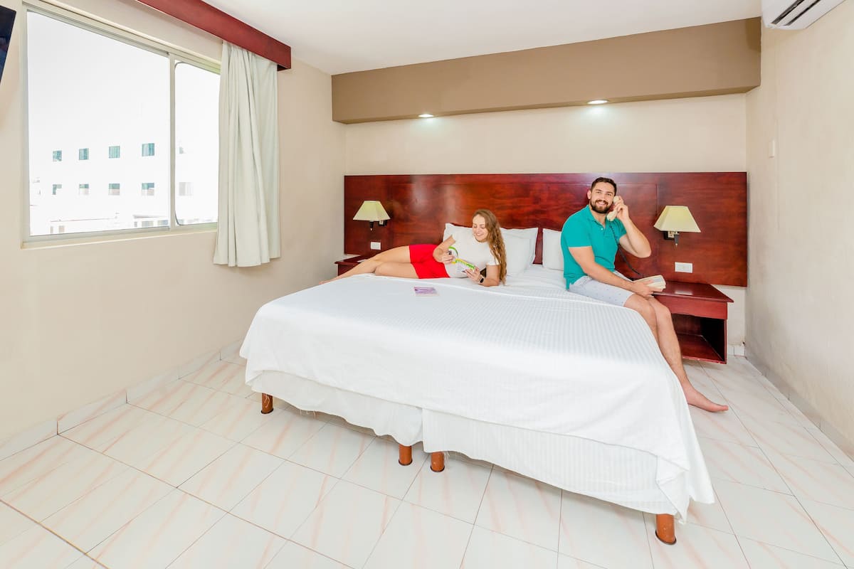A hotel room with a large bed, two bedside tables, a window with curtains, and a view of buildings outside.