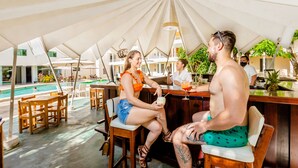 An outdoor bar area with wooden chairs and tables, a pool in the background, and umbrellas providing shade. - Hotel Maria del Carmen Merida (Mérida)