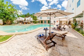 A pool area with lounge chairs and umbrellas, surrounded by greenery and a building with multiple windows. - Hotel Maria del Carmen Merida (Mérida)