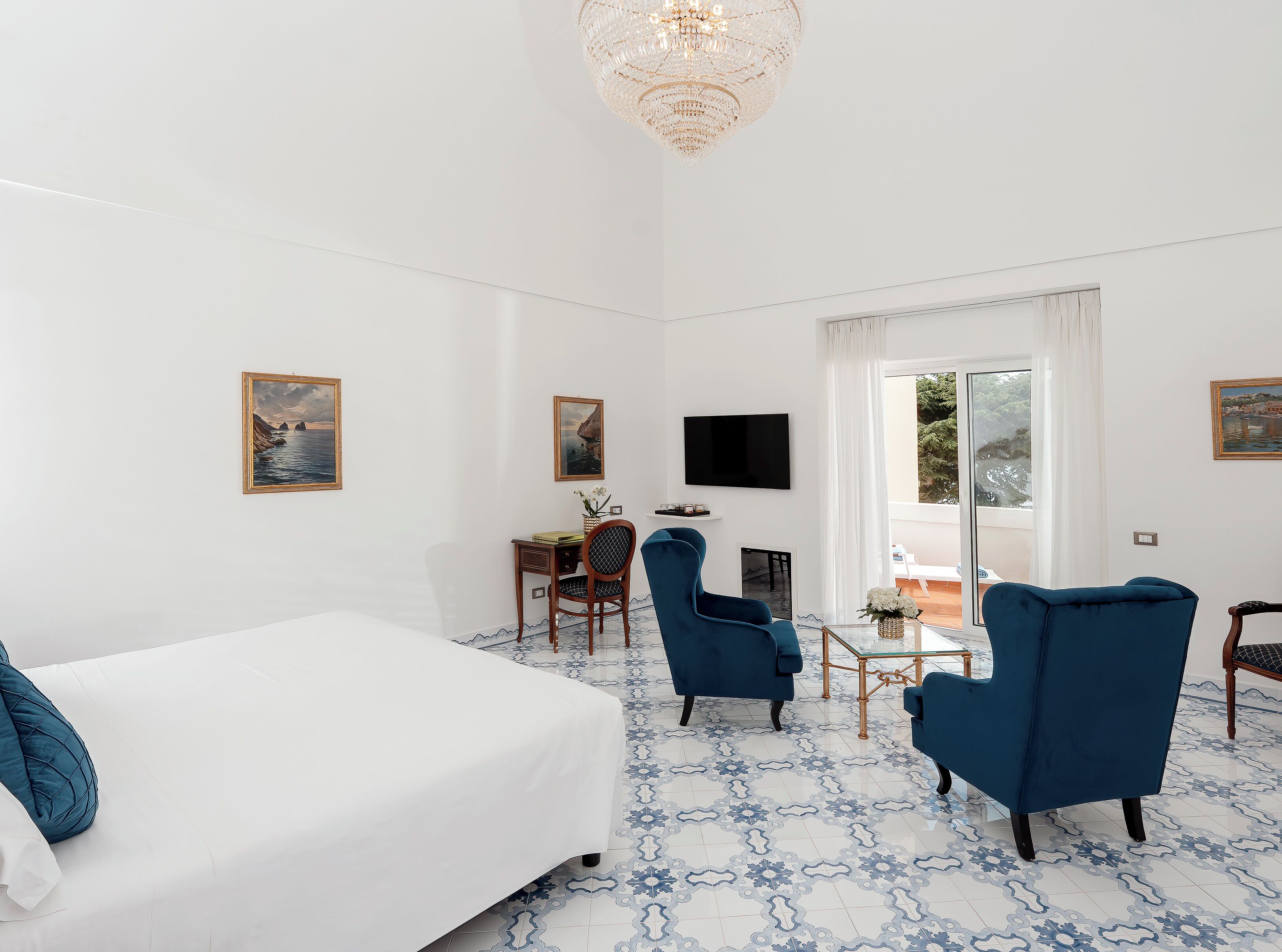 A hotel room with a large bed, two blue armchairs, a desk with a TV, and a large window offering a view of the outdoors.