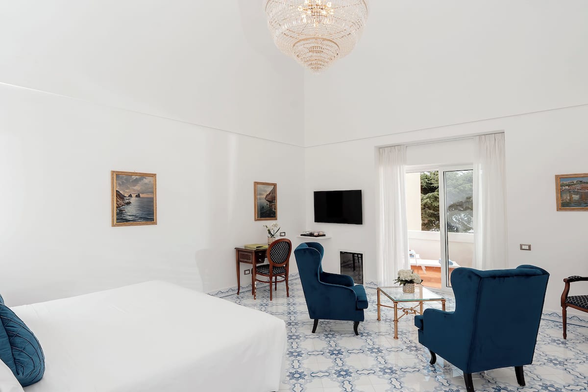 A hotel room with a large bed, two blue armchairs, a desk with a TV, and a large window offering a view of the outdoors.