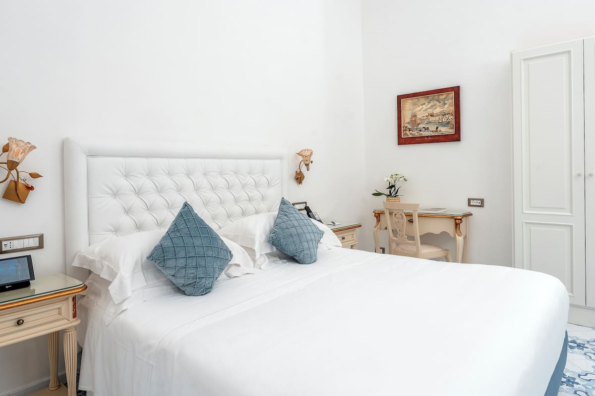 A neatly arranged bedroom with a large bed, a bedside table with a lamp, a small desk, and a chair.