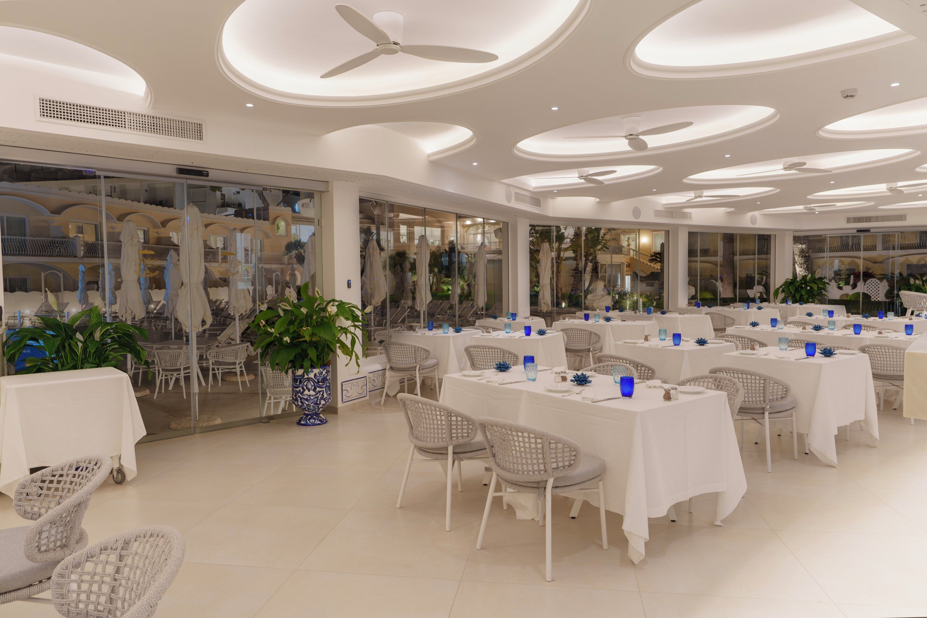A spacious indoor dining area with multiple tables and chairs, ceiling fans, and potted plants.