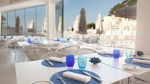 A modern dining area with white tables and chairs, blue glassware, and a view of a pool and umbrellas.