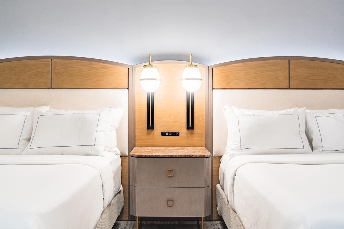 A hotel room with two beds, a wooden headboard, bedside tables with lamps, and a control panel above the headboard.