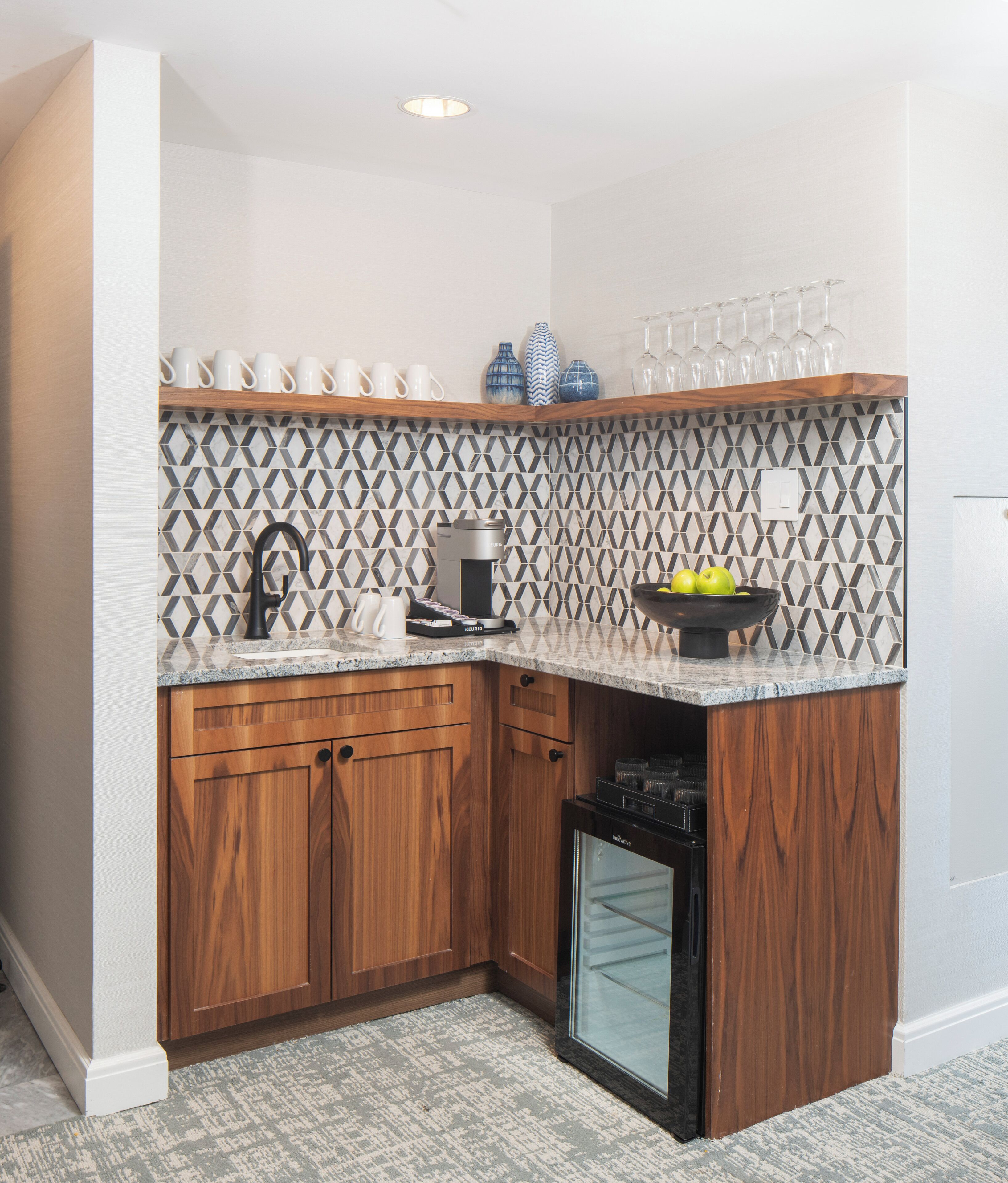 A compact kitchen with wooden cabinets, a built-in coffee maker, a small refrigerator, and a decorative backsplash.