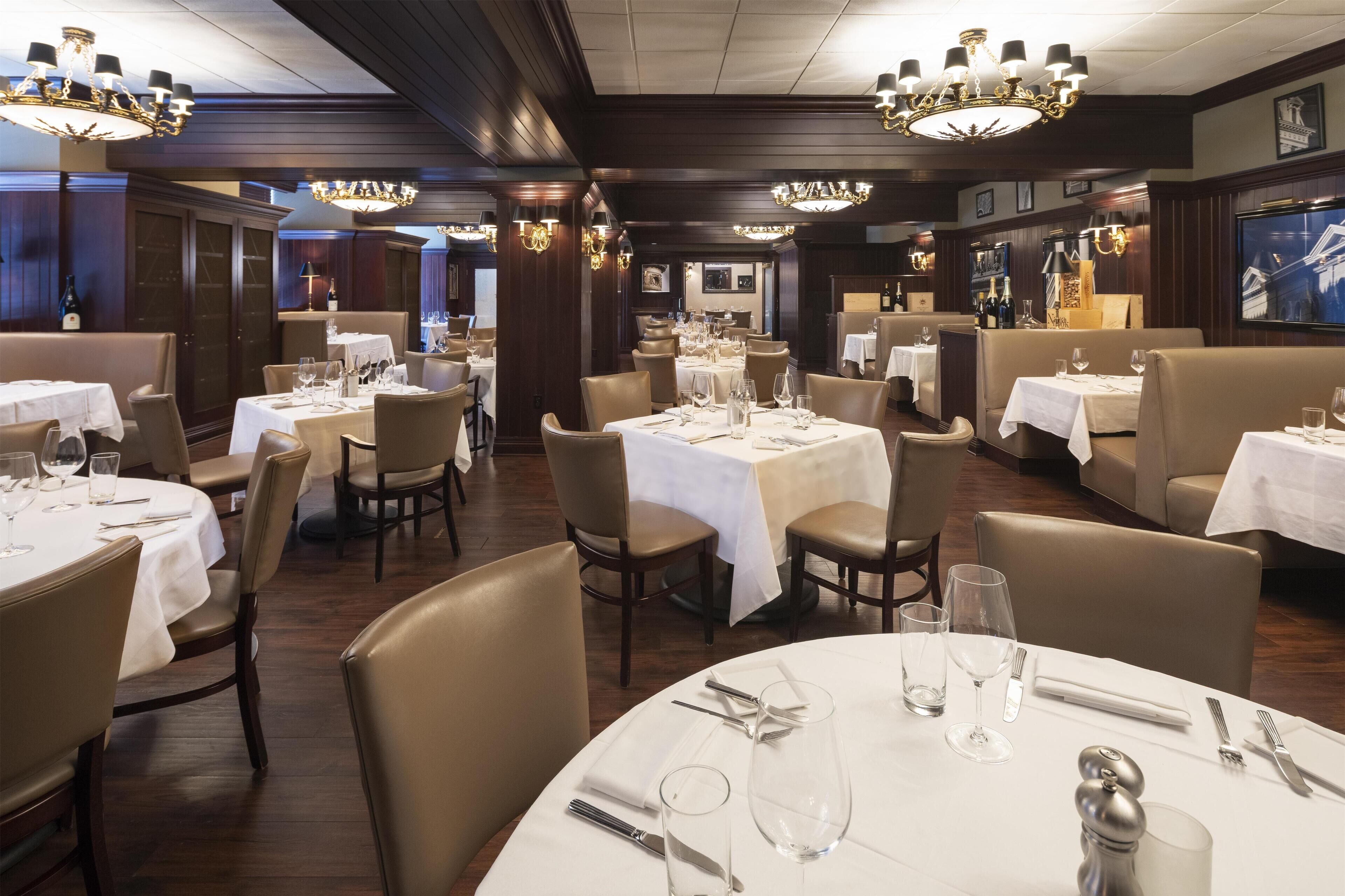 A restaurant with round tables set for dining, featuring chandeliers and wooden paneling.