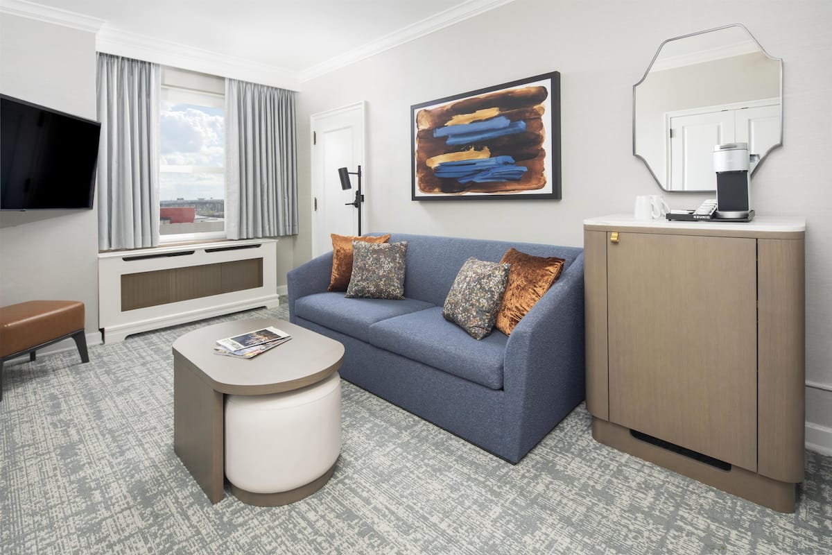 A hotel room with a sofa, ottoman, coffee machine, and a TV mounted on the wall.