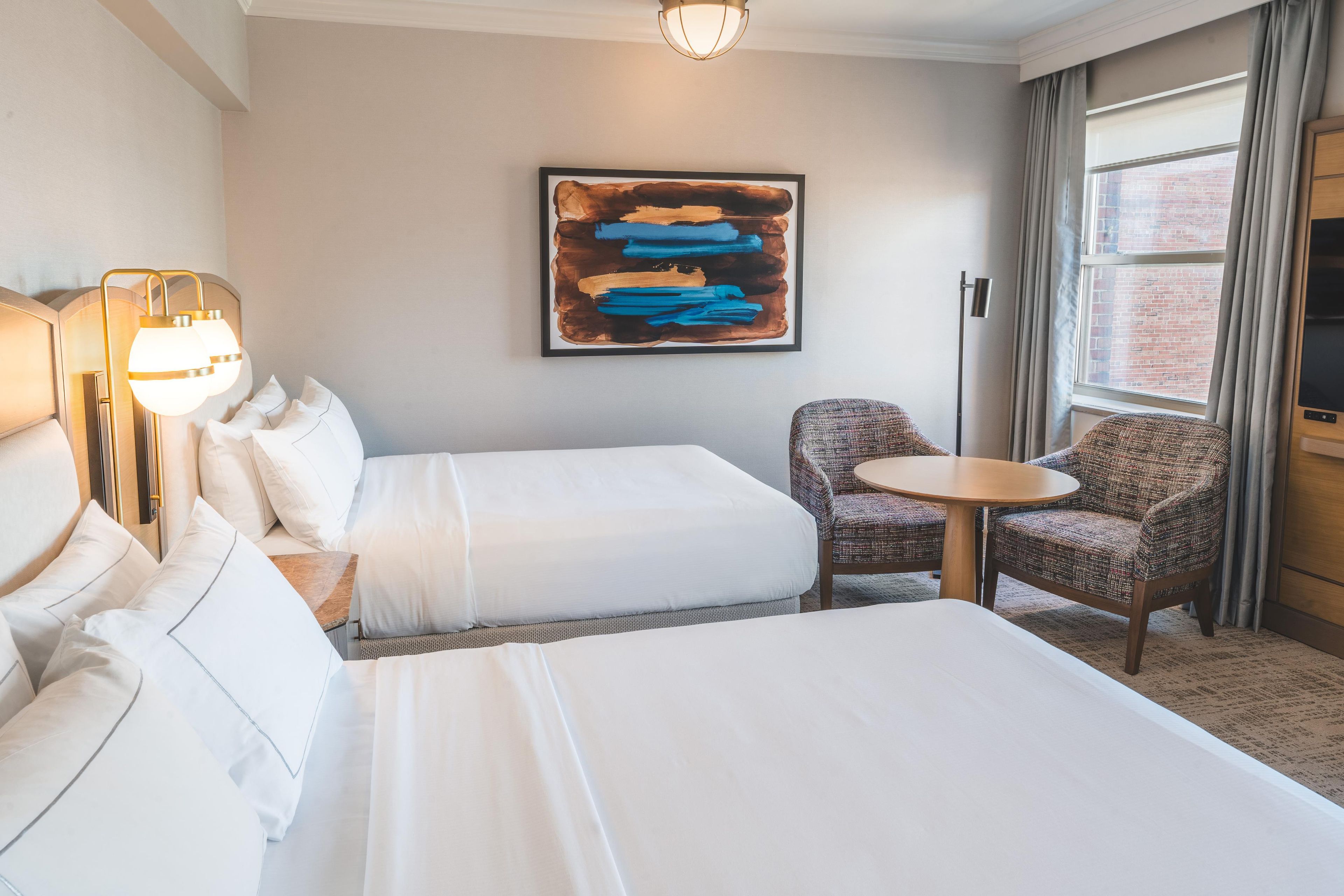 A hotel room with two beds, a small round table, a chair, and a painting on the wall.