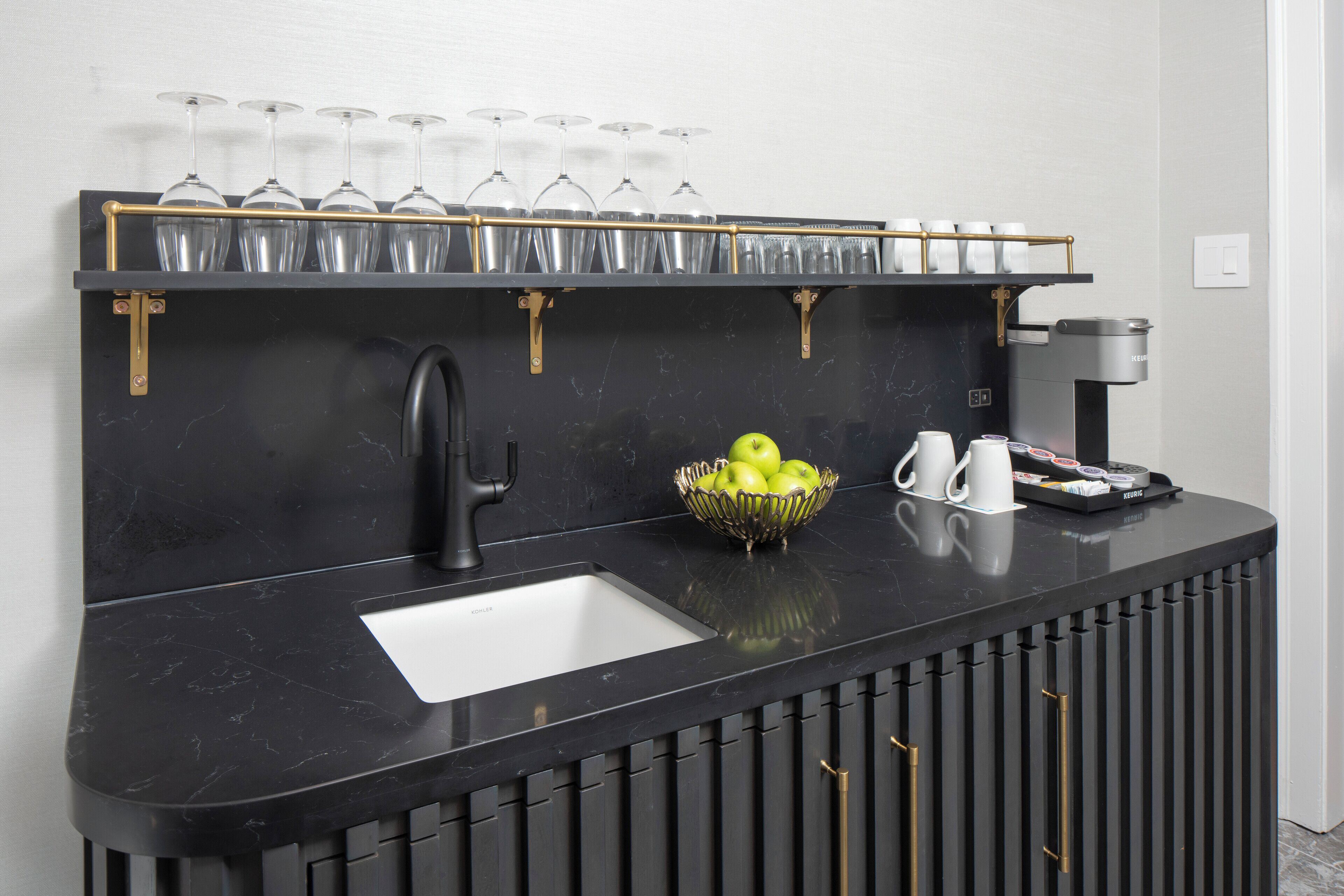 A black marble bar with a sink, a coffee machine, a bowl of green apples, and a wine glass rack.