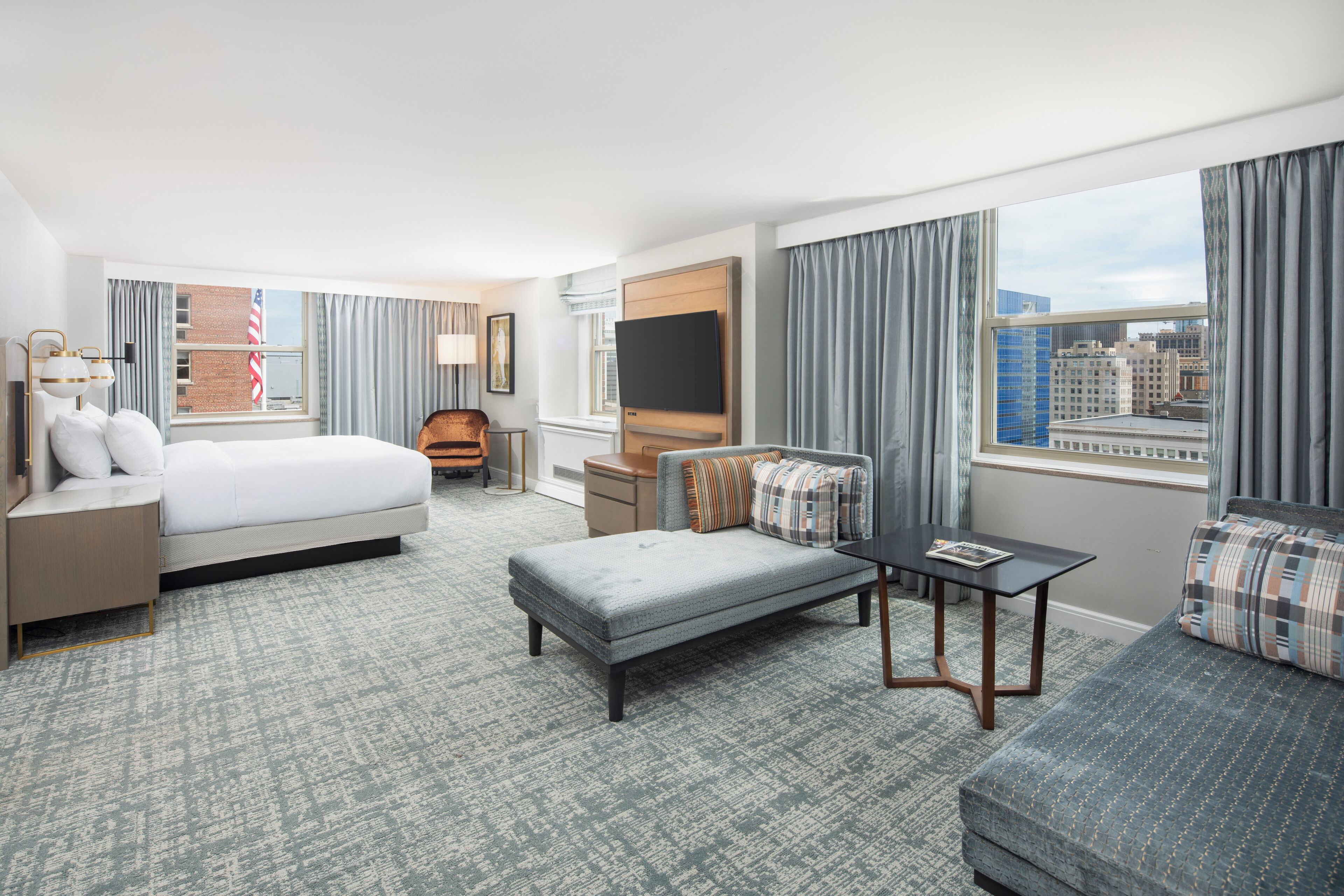 A hotel room with a large bed, a sofa, a small table, a TV, and a window with a city view.