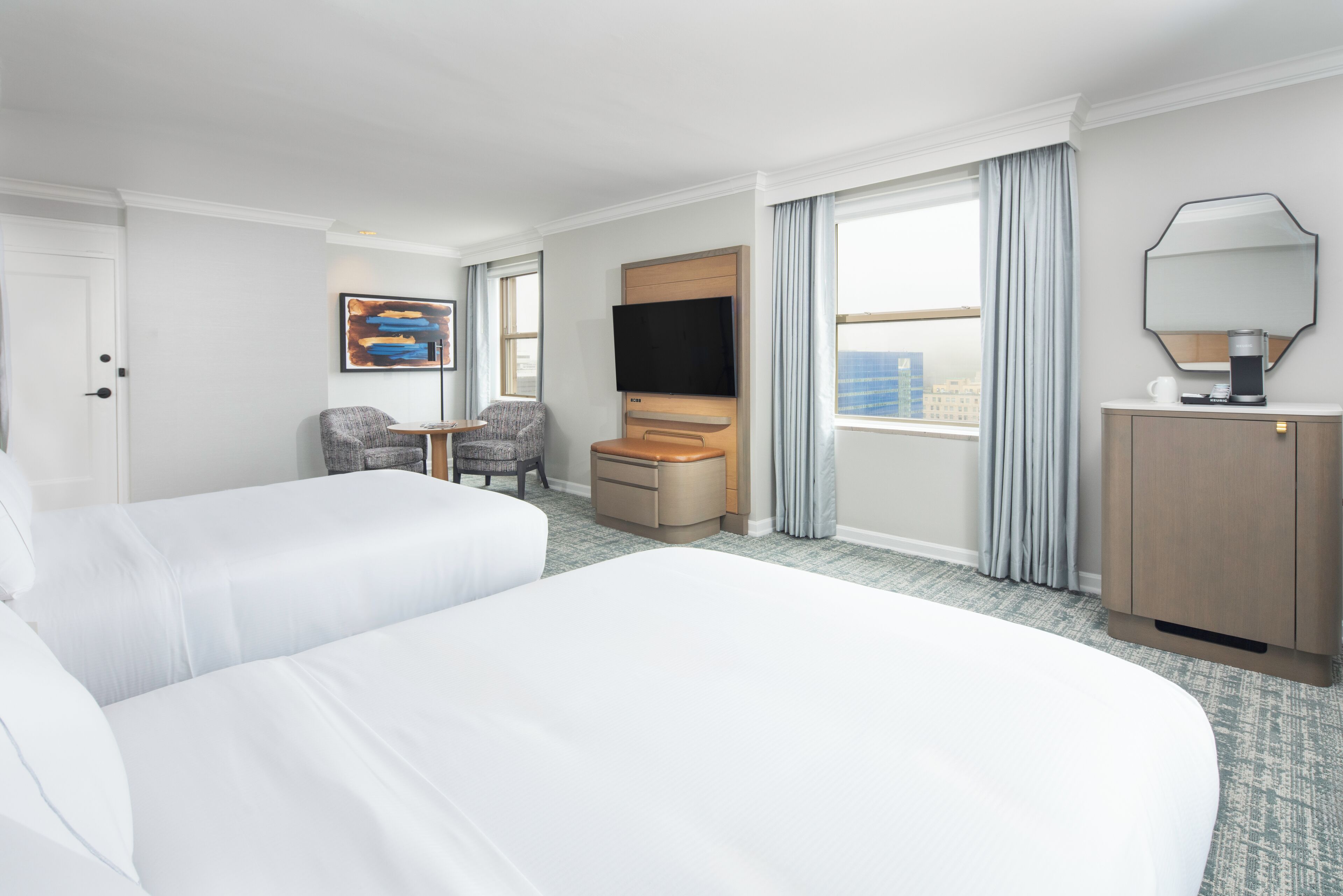 A hotel room with two beds, a TV, a desk, a chair, and a large window with curtains.