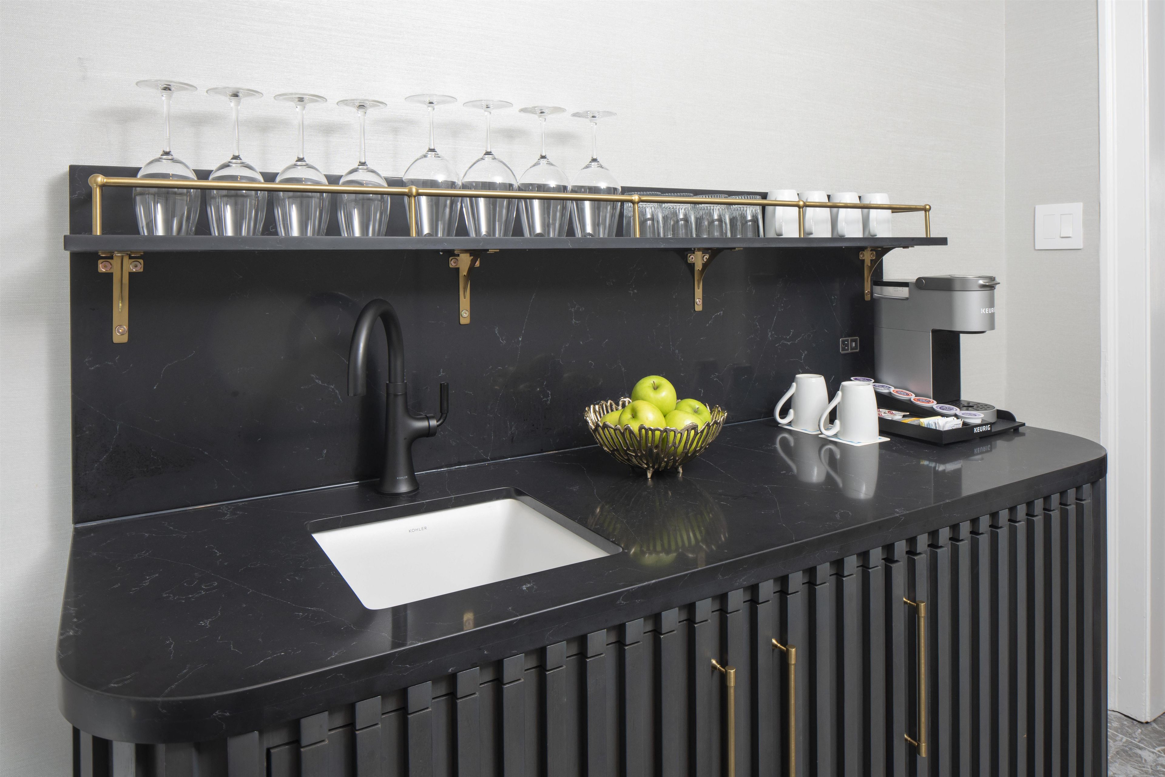 A black marble bar with a sink, a coffee machine, a bowl of apples, and a wine glass rack.