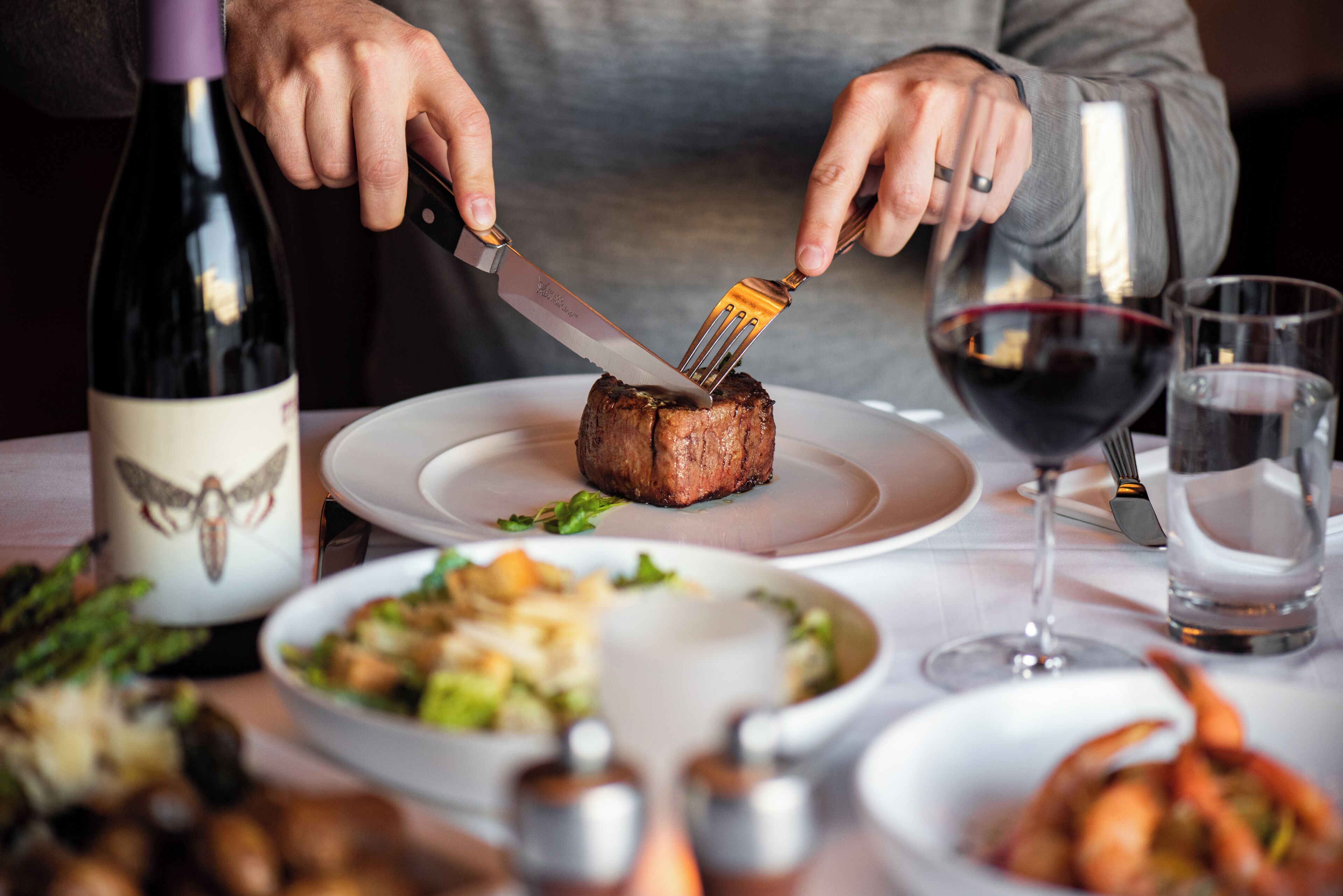 Eine Person schneidet ein Steak mit Messer und Gabel auf einem Teller, dazu steht eine Weinflasche und Gläser auf dem Tisch.