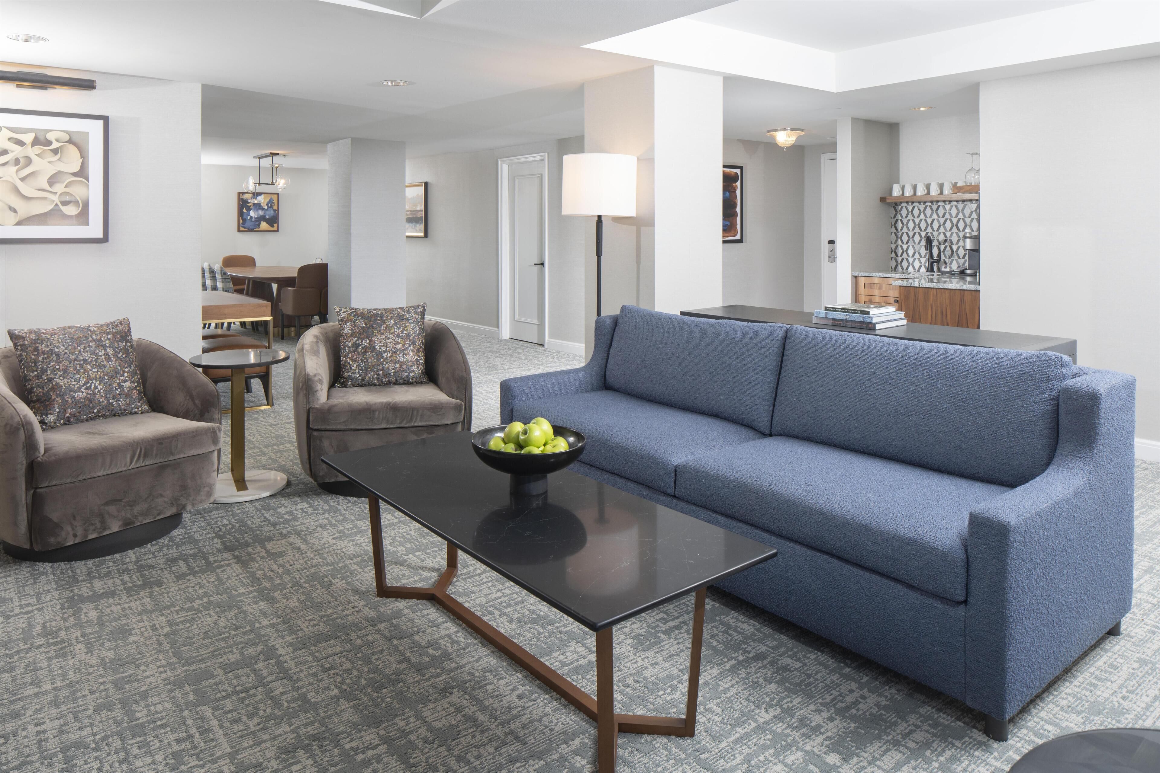 A modern living room with a blue sofa, two armchairs, a coffee table, and a dining area in the background.