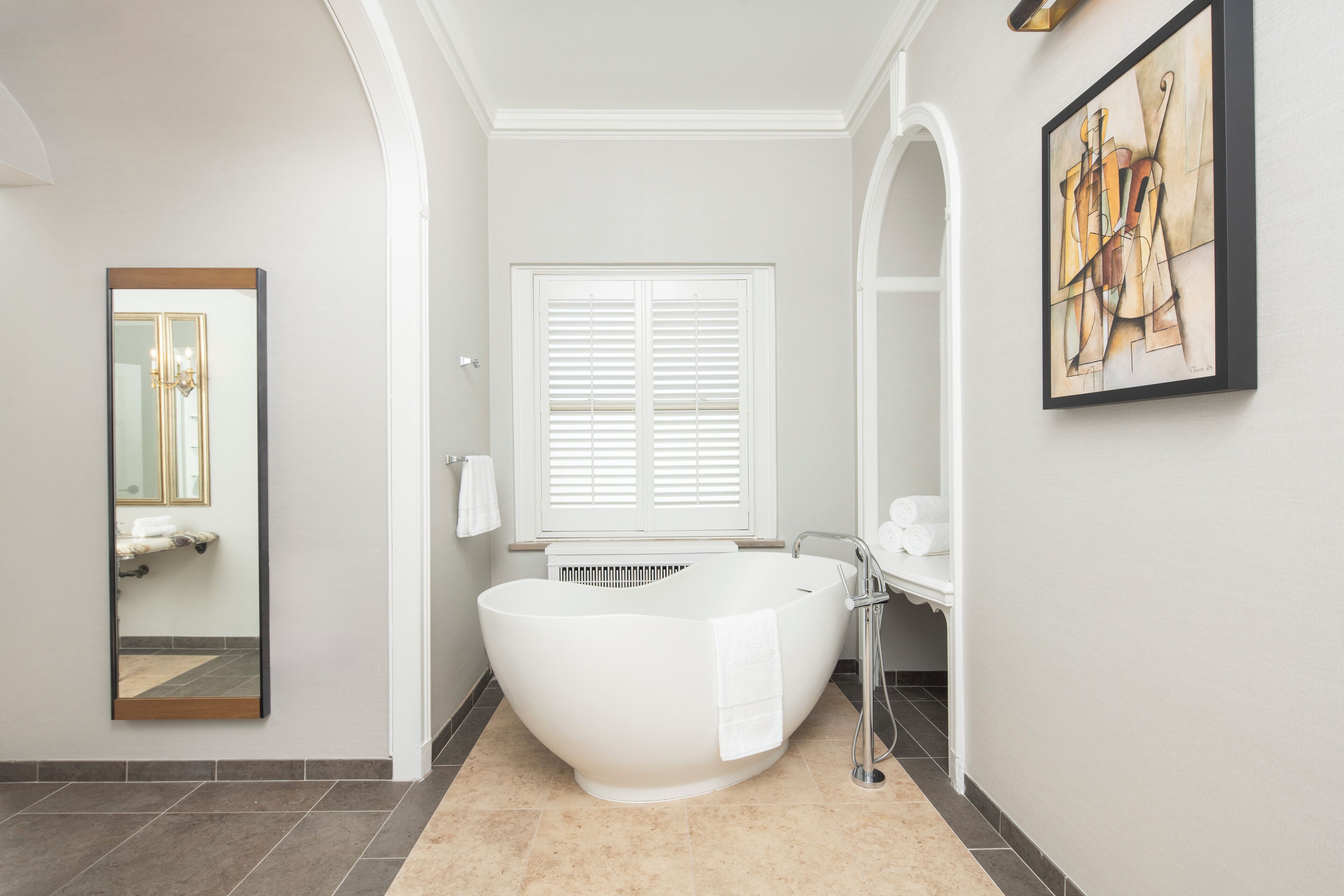 Ein modernes Badezimmer mit einer freistehenden Badewanne, einem großen Spiegel und einem Fenster mit Jalousien.