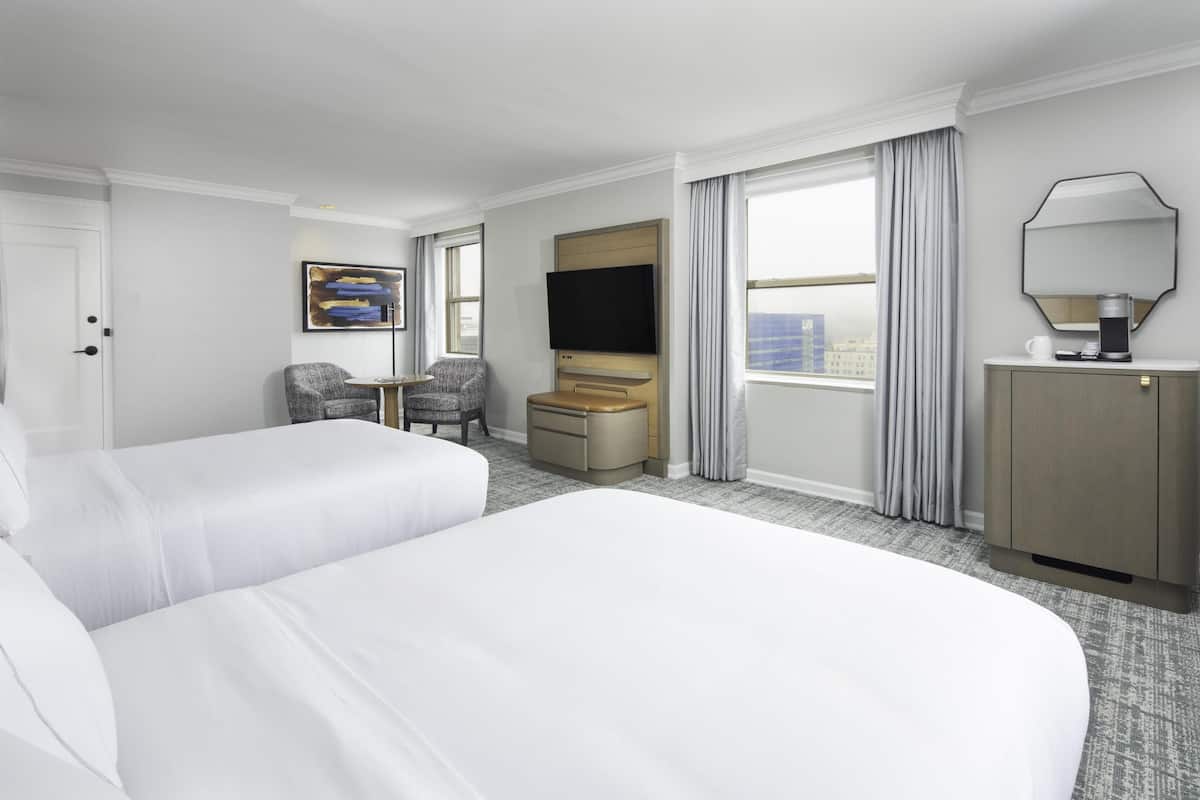 A hotel room with two beds, a TV, a desk, a chair, and a large window with curtains.