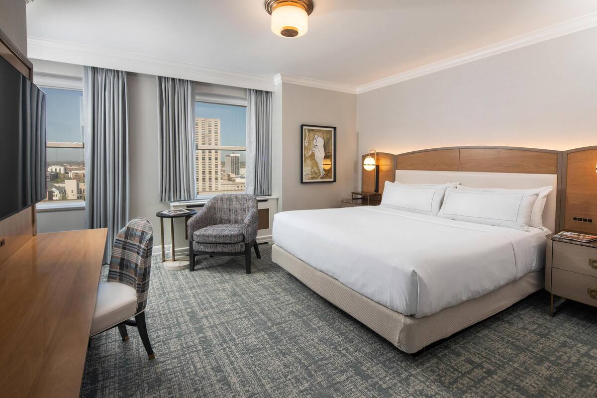 A hotel room with a large bed, a desk, a chair, a nightstand, and a view of the city through the window.