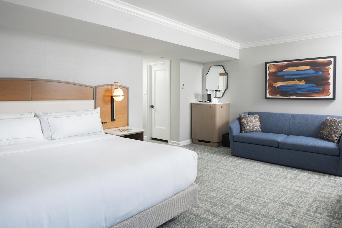 A hotel room with a large bed, a sofa, a nightstand with a lamp, a dresser, and a framed abstract painting on the wall.