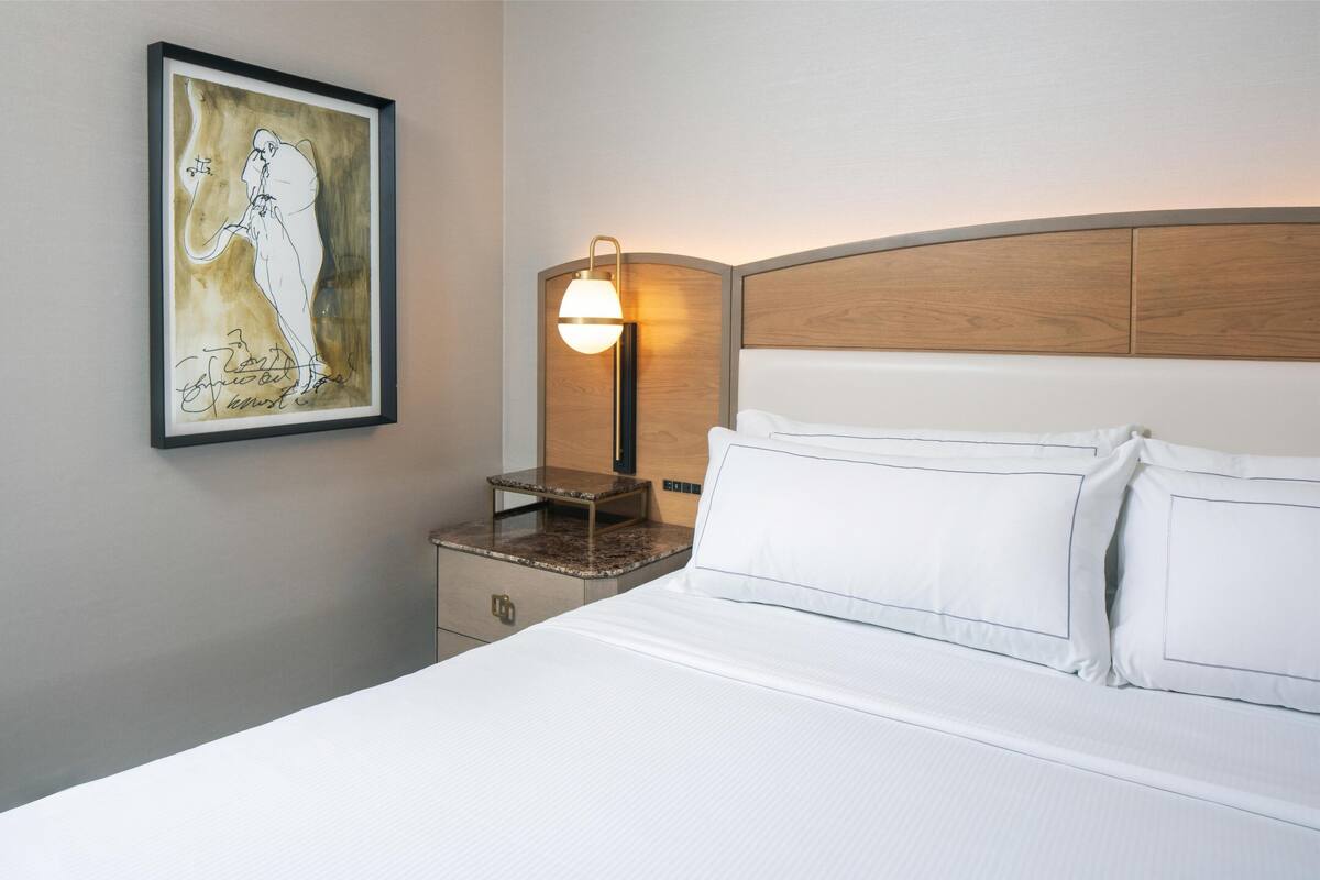 A hotel room with a bed, a nightstand, a framed artwork, and a wall-mounted lamp.
