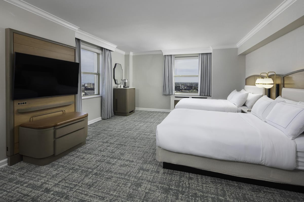 A hotel room with two beds, a flat-screen TV, a dresser, a desk, and a large window offering a city view.