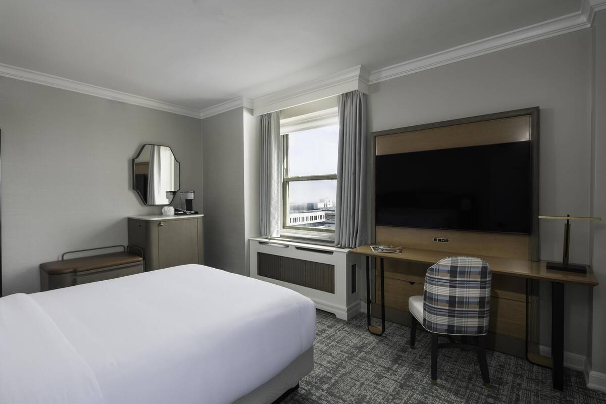 A hotel room with a large bed, a desk with a chair, a flat-screen TV, a window with curtains, a mirror, and a radiator.