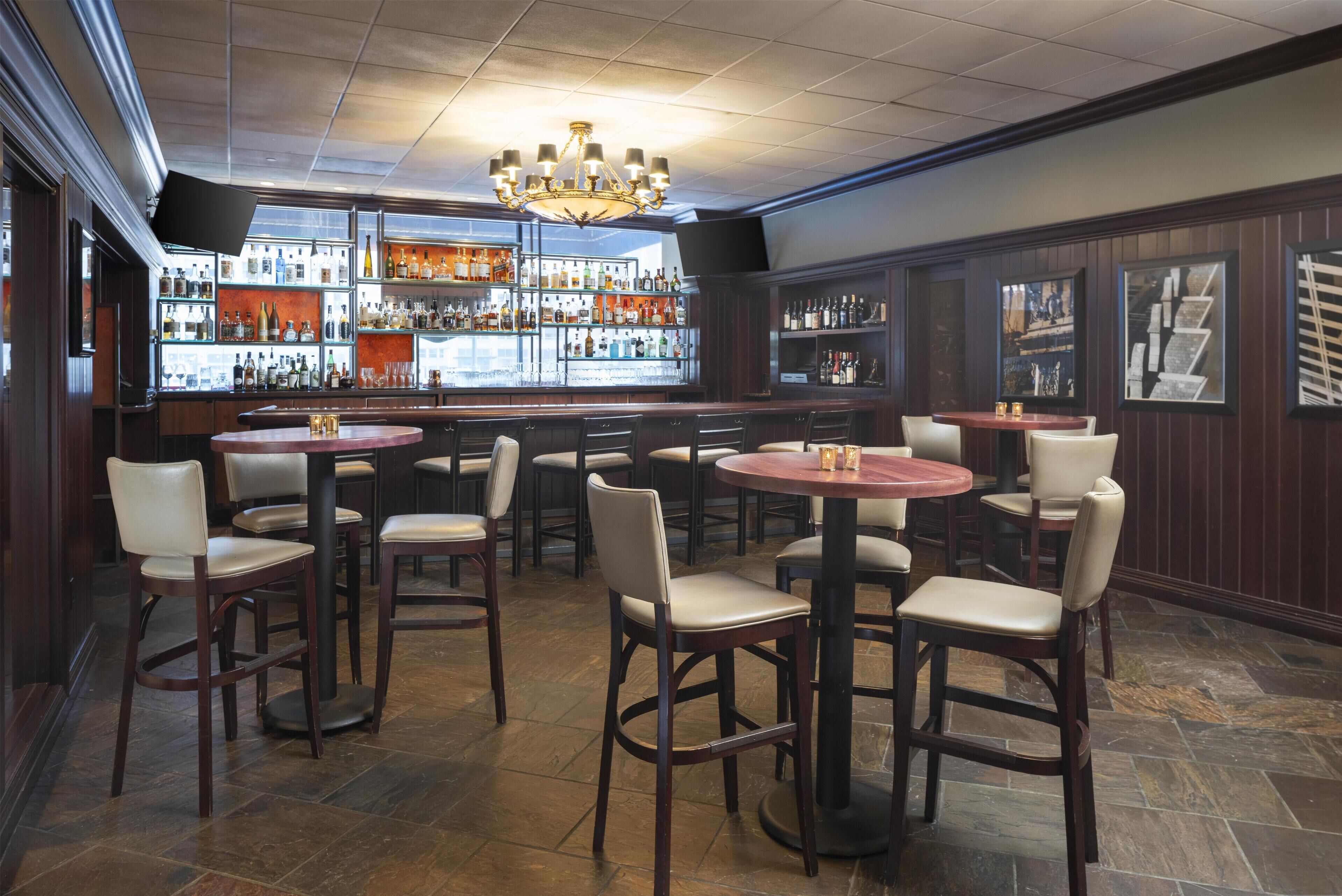 A bar area with multiple round tables and chairs, a chandelier, and a well-stocked bar counter.