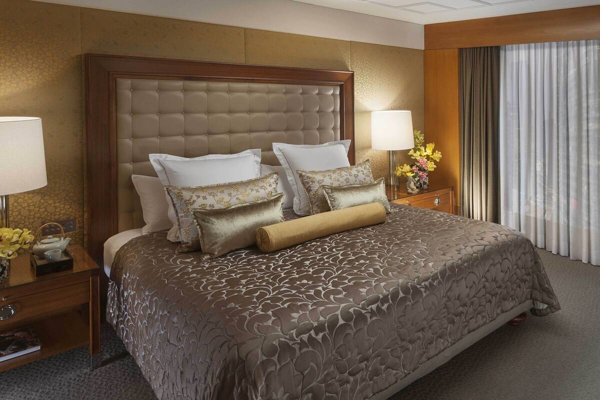 A large bed with a tufted headboard, multiple pillows, and a decorative throw. There is a nightstand with a lamp and a vase of flowers. The room has a carpeted floor and a window with curtains.