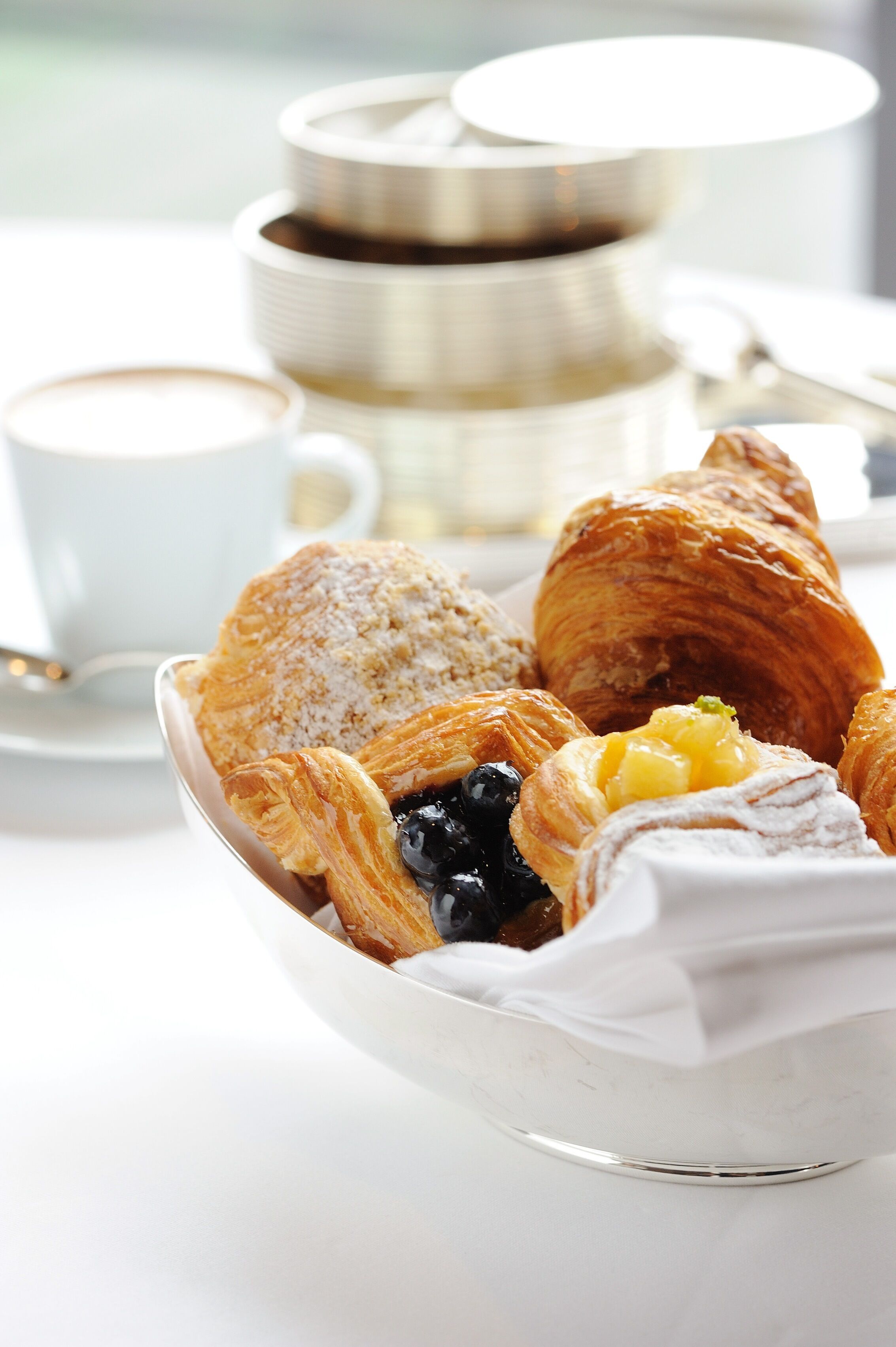 A tray of pastries including croissants and a dessert with blueberries and whipped cream, accompanied by a cup of coffee and a teapot in the background.