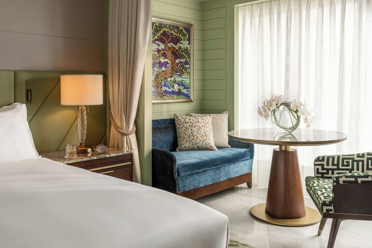 A hotel room with a bed, a nightstand with a lamp, a blue sofa, a round table with a vase of flowers, and a painting on the wall.
