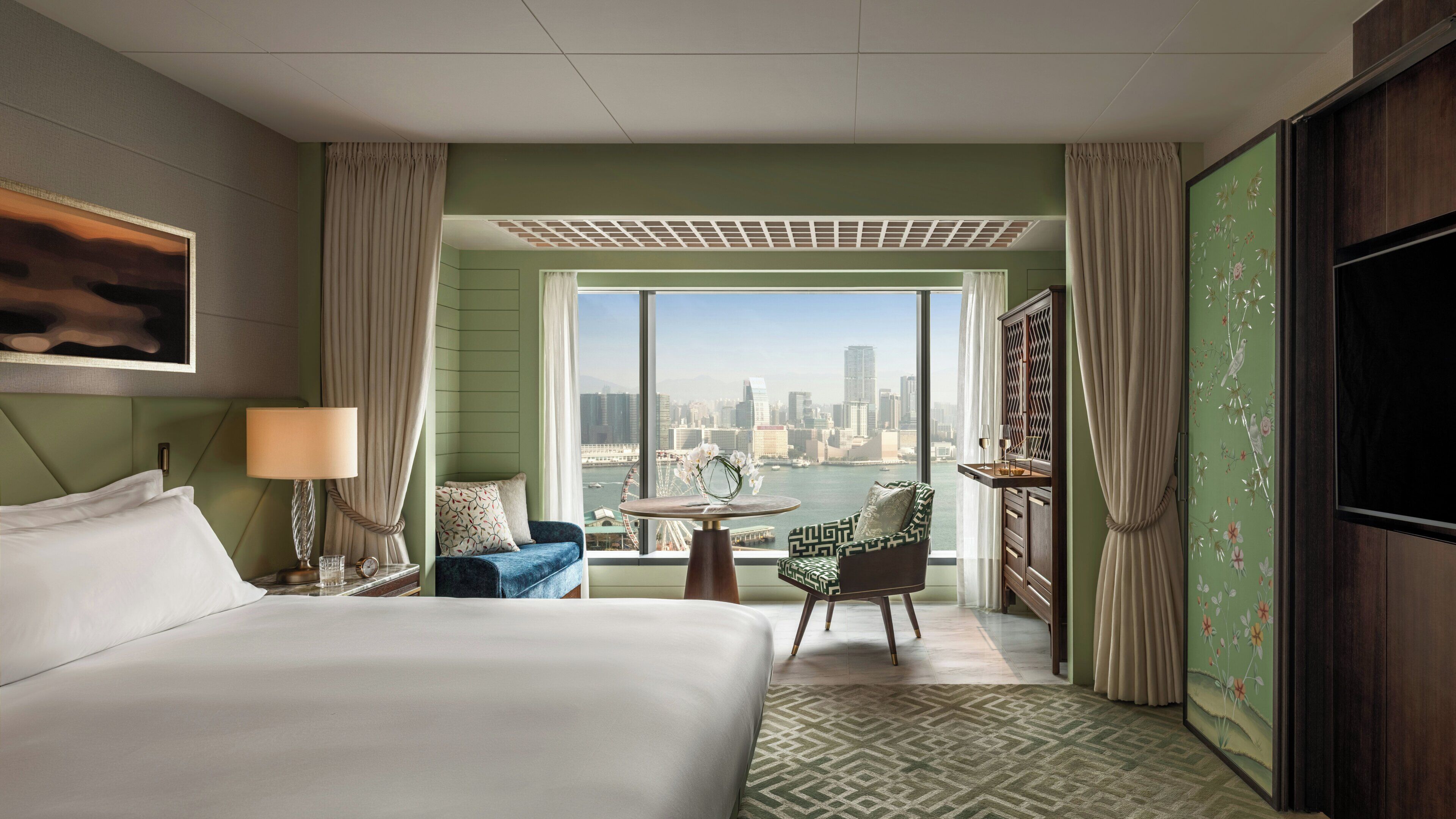 Habitación de hotel con cama, zona de estar con mesa, vista panorámica de la ciudad y un ventanal grande.