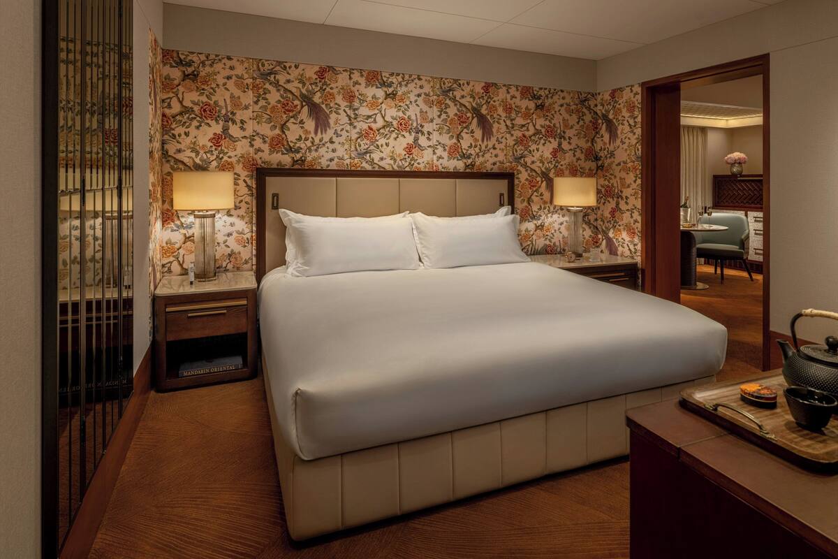 A hotel room with a large bed, two bedside tables with lamps, floral wallpaper, and a view into the adjacent room.