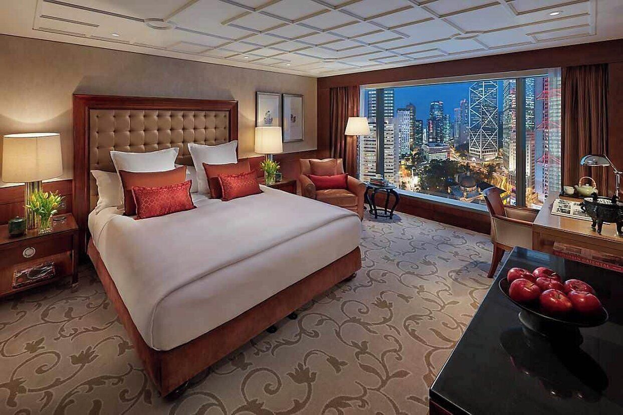 A hotel room with a large bed, two chairs, a table with apples, and a city view.