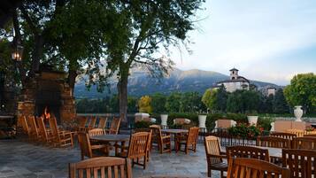 An outdoor patio with wooden tables and chairs, a stone fireplace, and a view of a mountain and a building in the background.