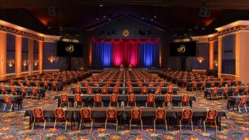 Un salón de conferencias con filas de mesas y sillas, un escenario con cortinas y una pantalla grande.