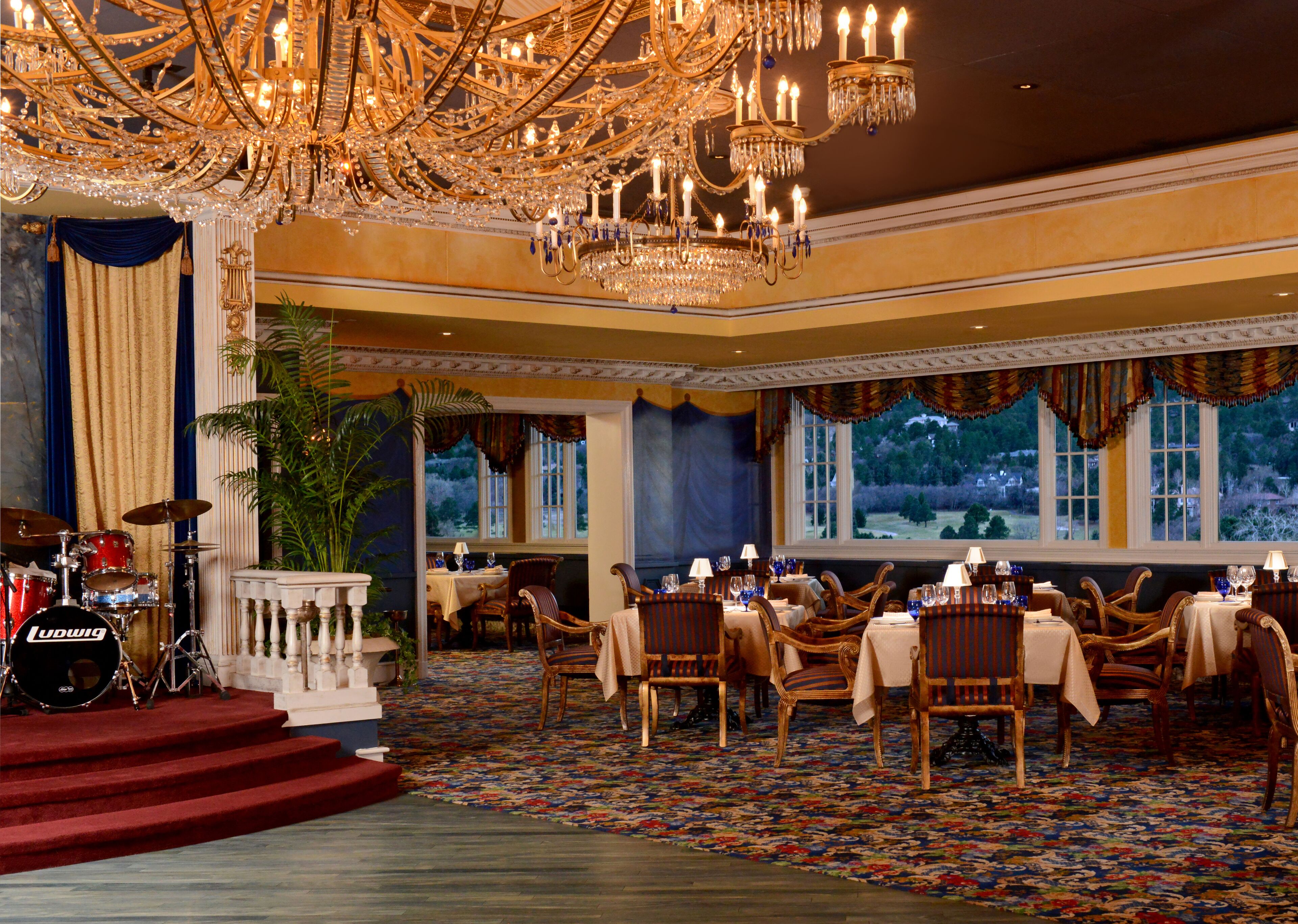 A grand dining room with ornate chandeliers, a stage with a drum set, and a scenic view through large windows.