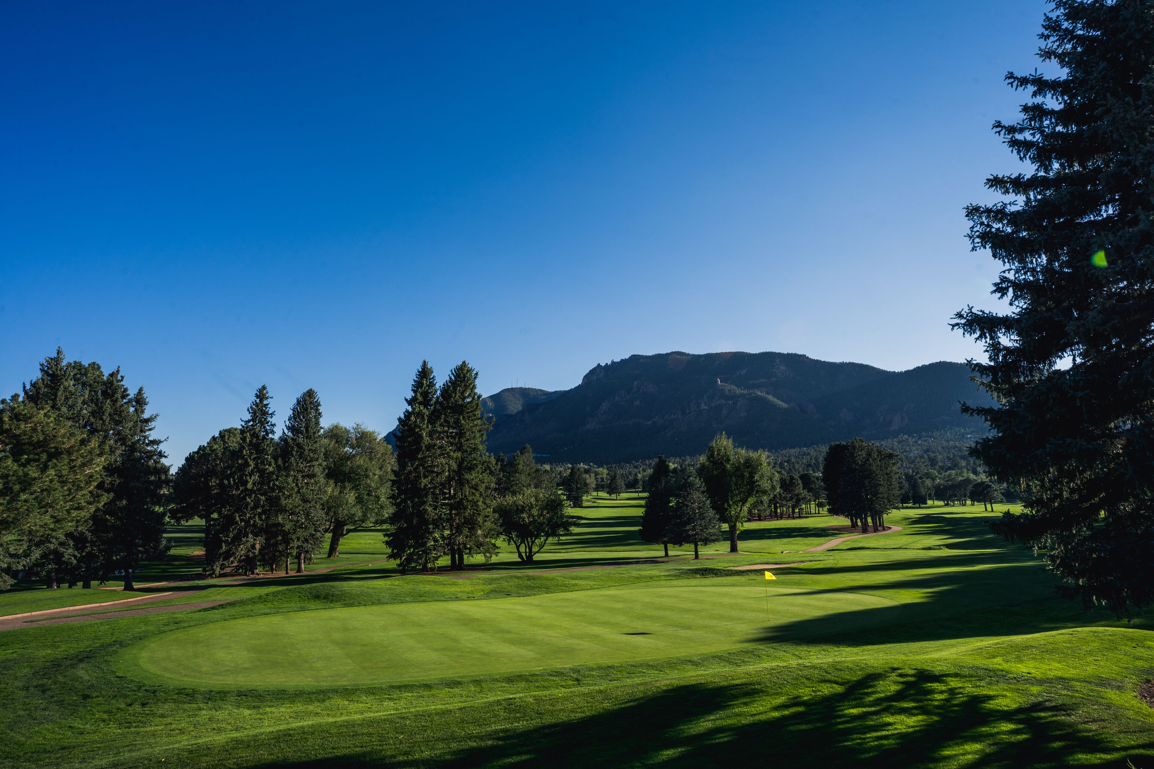 Un campo de golf con cielo despejado, árboles y montañas al fondo.
