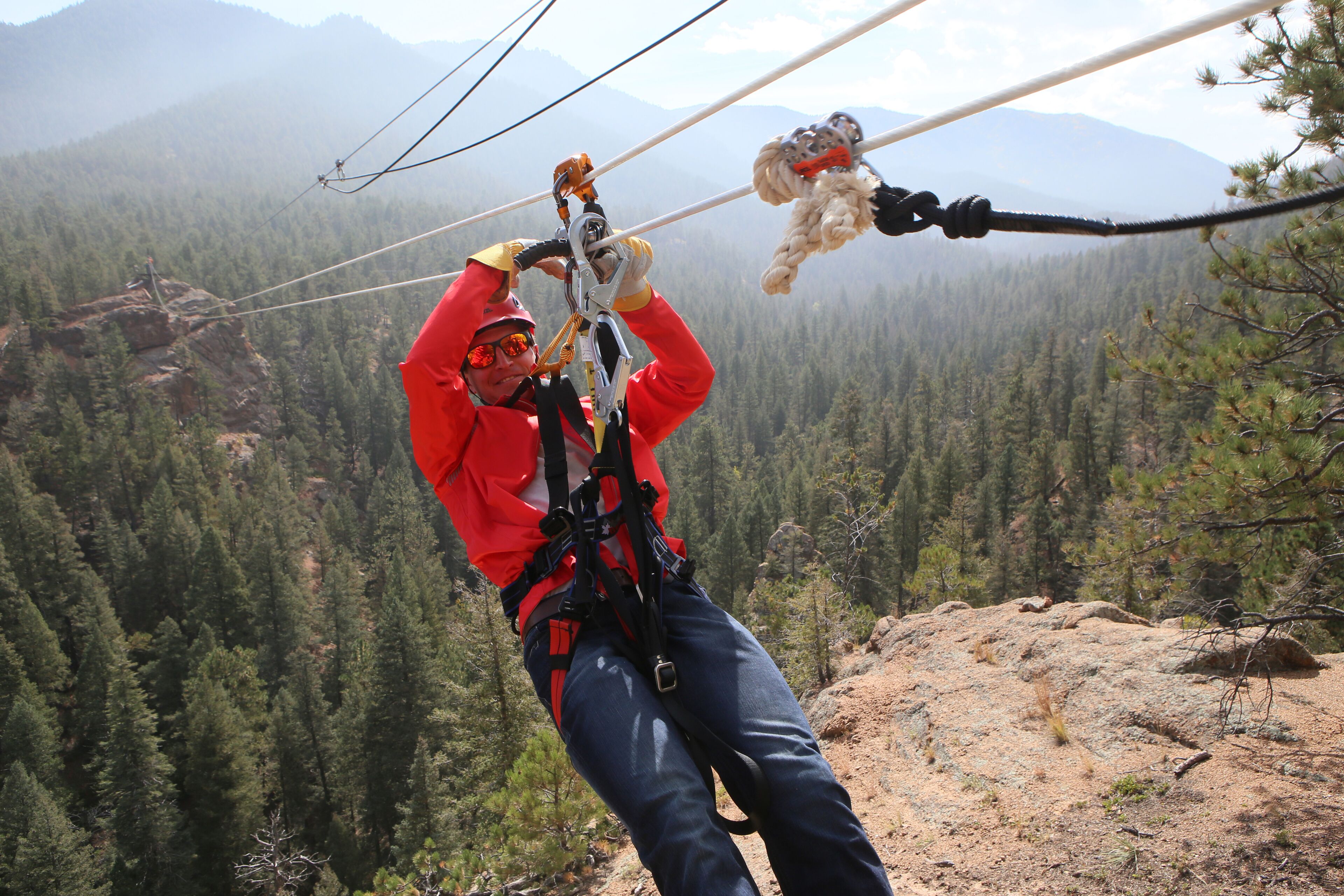 A person in a red jacket and blue pants engaged in a zip-lining activity, with a forested mountain landscape in the background.