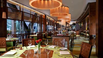A restaurant with wooden tables, green cushioned chairs, and a large window with a view of trees.