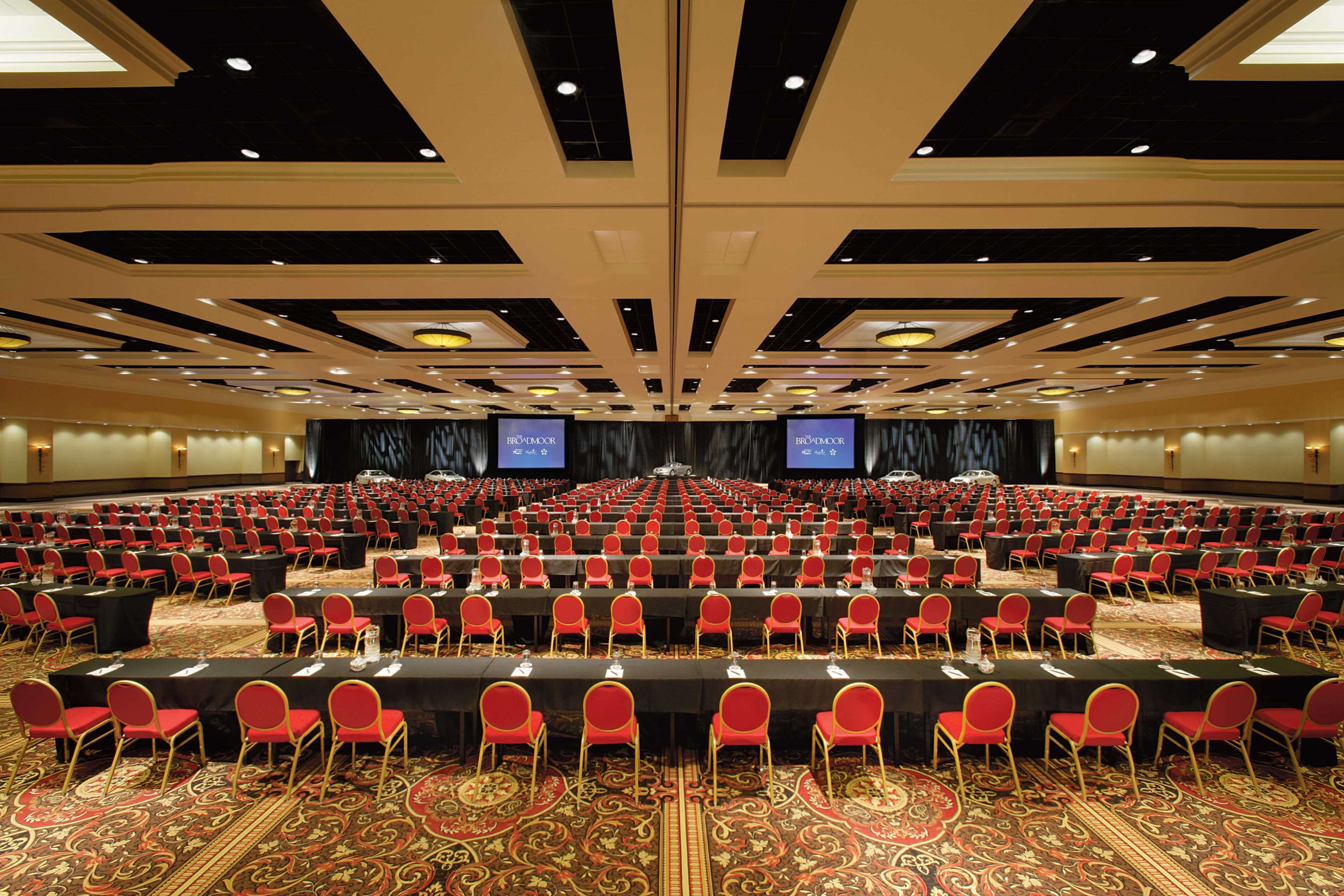 A large conference room with rows of red chairs and tables, a stage with a screen, and a carpeted floor.
