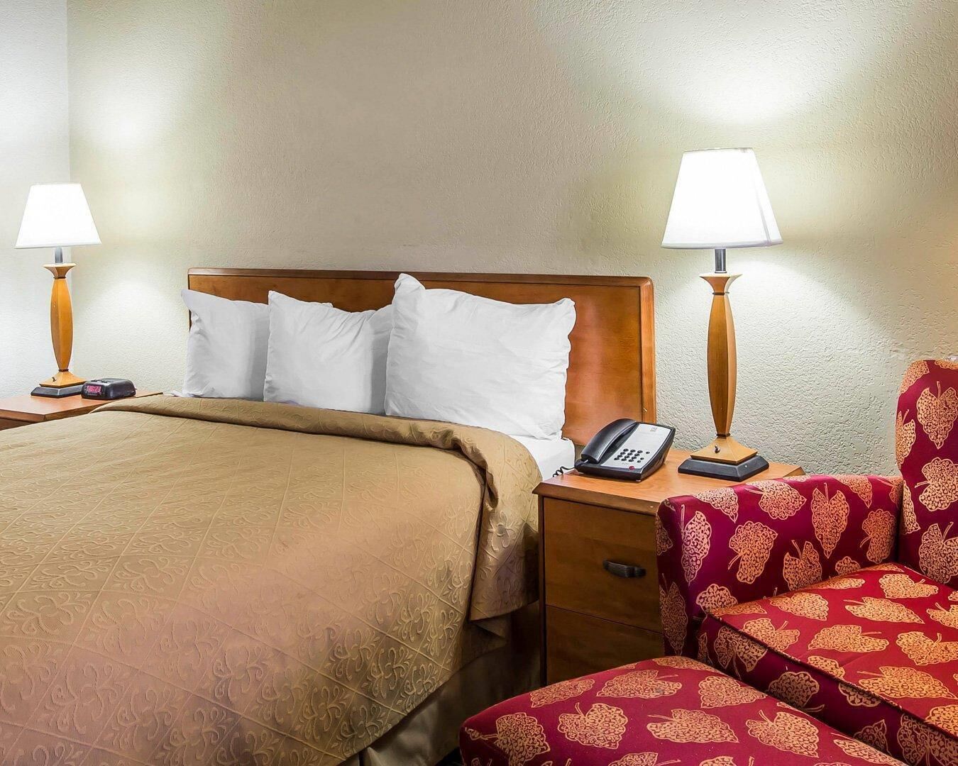 A hotel room with a bed, two bedside lamps, a telephone, a chair, and a nightstand.