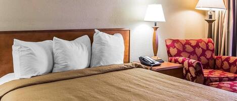 A hotel room with a bed, pillows, a bedside table with a lamp, a telephone, and a patterned armchair.
