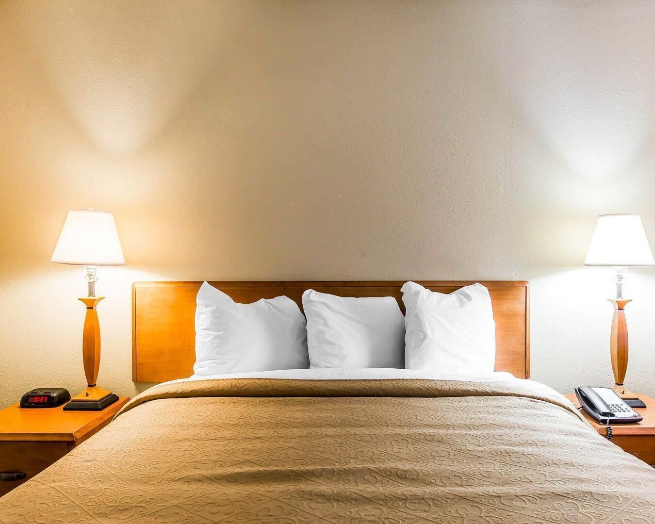 A hotel room with a bed, two bedside lamps, a nightstand with a phone, and a wall clock.
