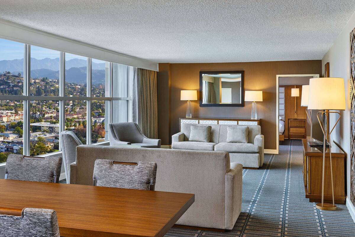 Hotelkamer met groot raam en uitzicht op de stad, een bank, een eettafel en een lamp.