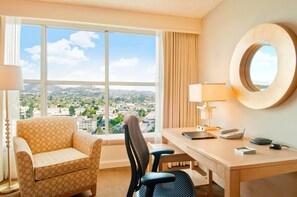A hotel room with a large window offering a city view, a desk with a lamp, a chair, and a round mirror on the wall.