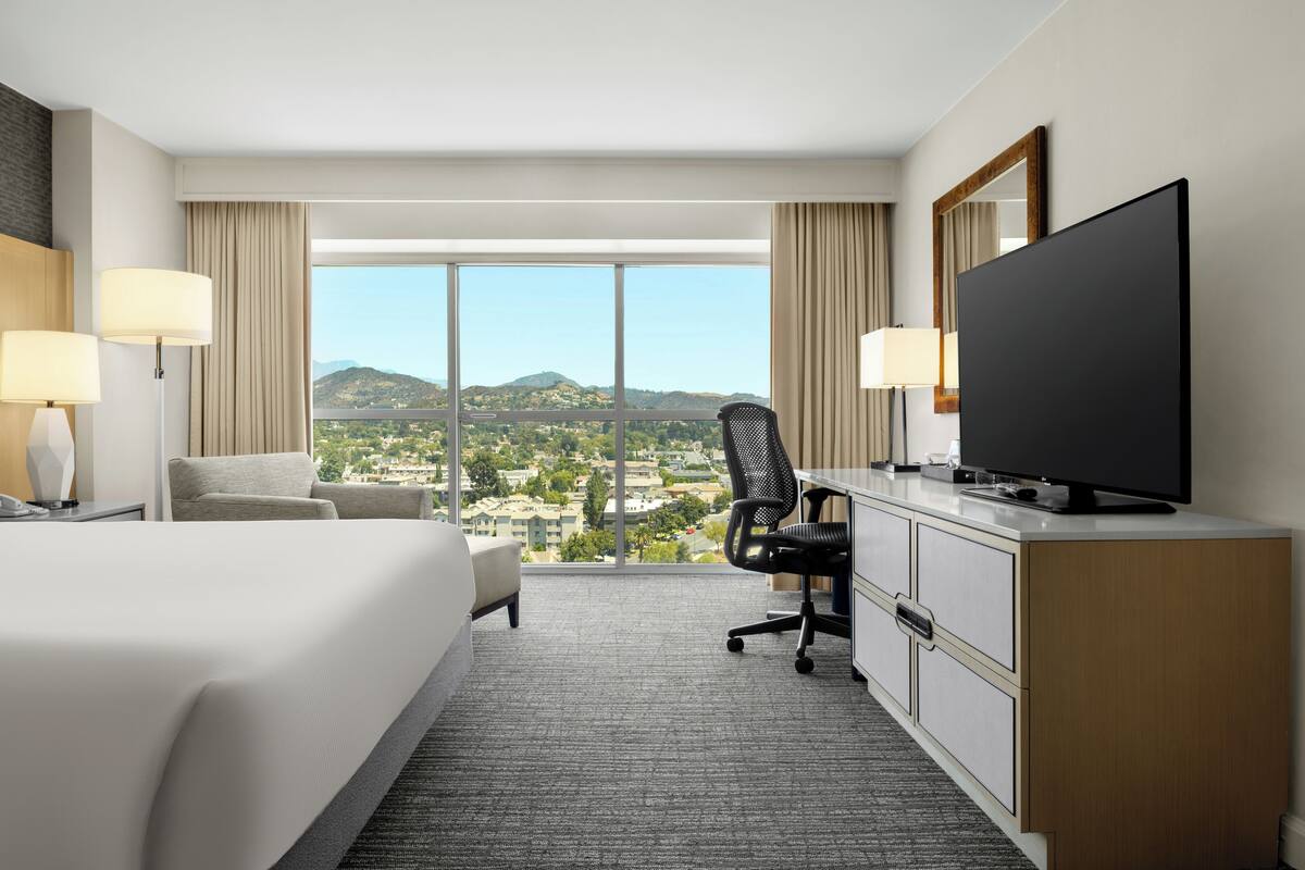 Een hotelkamer met een groot bed, een bureau met stoel, een televisie en uitzicht op een stadsgezicht door grote ramen.