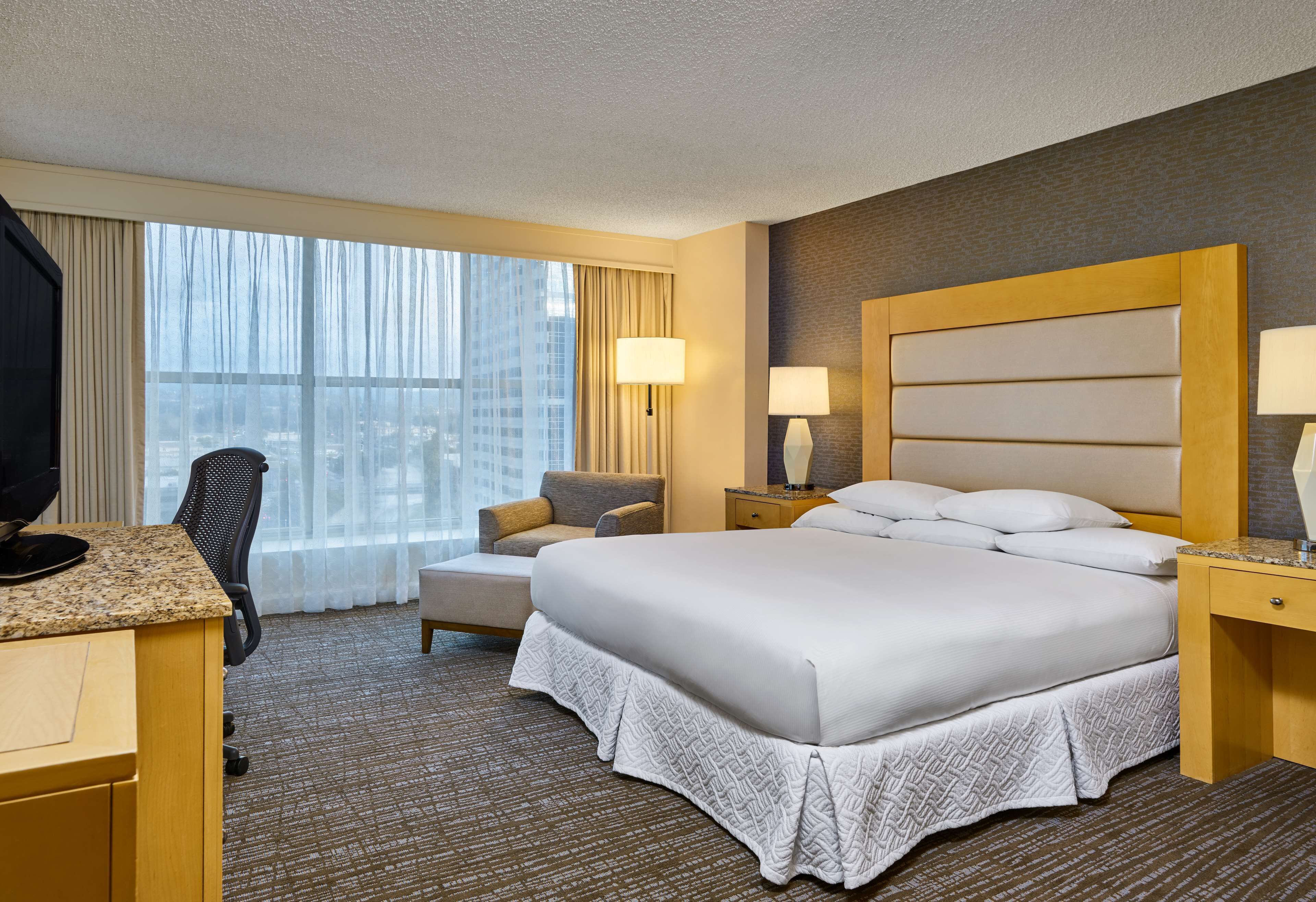 Una habitación de hotel con una cama grande, un escritorio con computadora, una silla, una mesita de noche con lámparas y vistas a la ciudad a través de amplios ventanales.