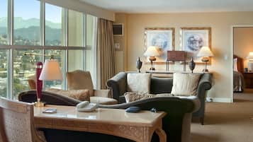 A hotel room with a large window, two framed pictures on the wall, a sofa, a table with a lamp, and a chair.
