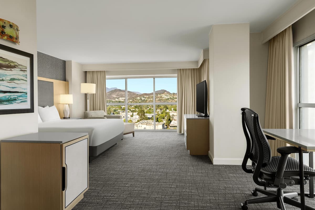 Hotelkamer met een bed, bureau, stoel, televisie en een groot raam met uitzicht op bergen.